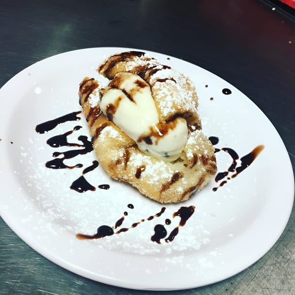 Fried dough dessert with vanilla ice cream, chocolate drizzle, and powdered sugar on a white plate.